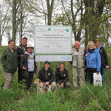 People dressed in outdoor wear standing beside middle point woods western lake erie islands national area sign dispenser amenities