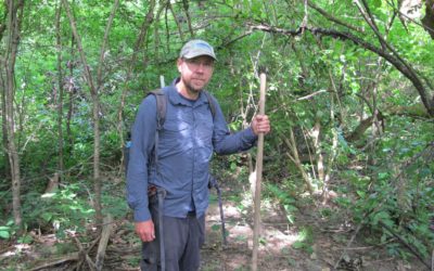 man standing in woods holding wooden stick wearing backpack dispenser amenities