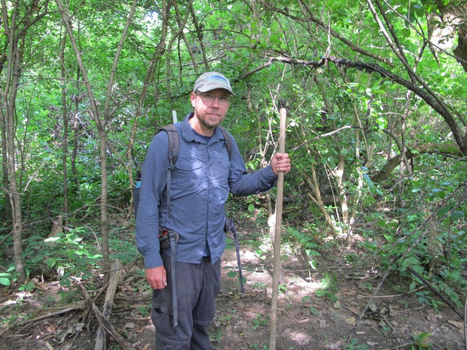 Man standing in woods holding wooden stick wearing backpack dispenser amenities